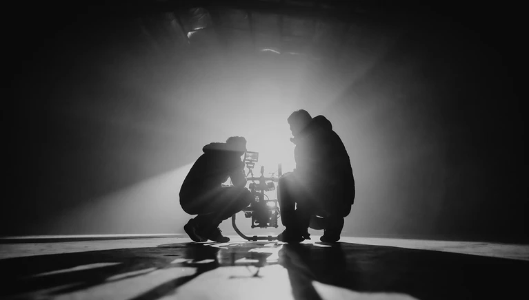 Two people silhouetted under spotlight appearing to collaborate or assist each other.