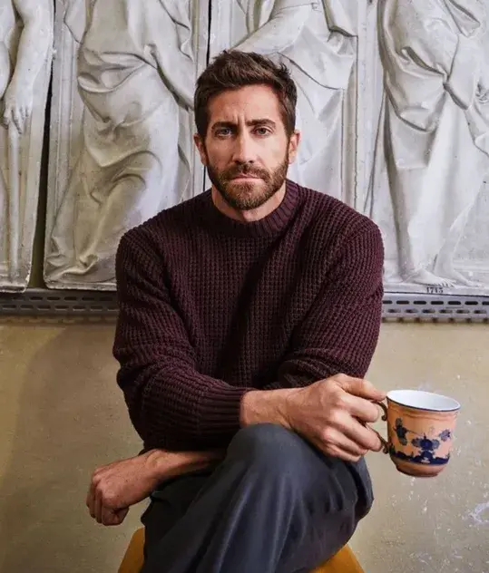 Man in maroon sweater holding a floral mug, seated before stone reliefs.